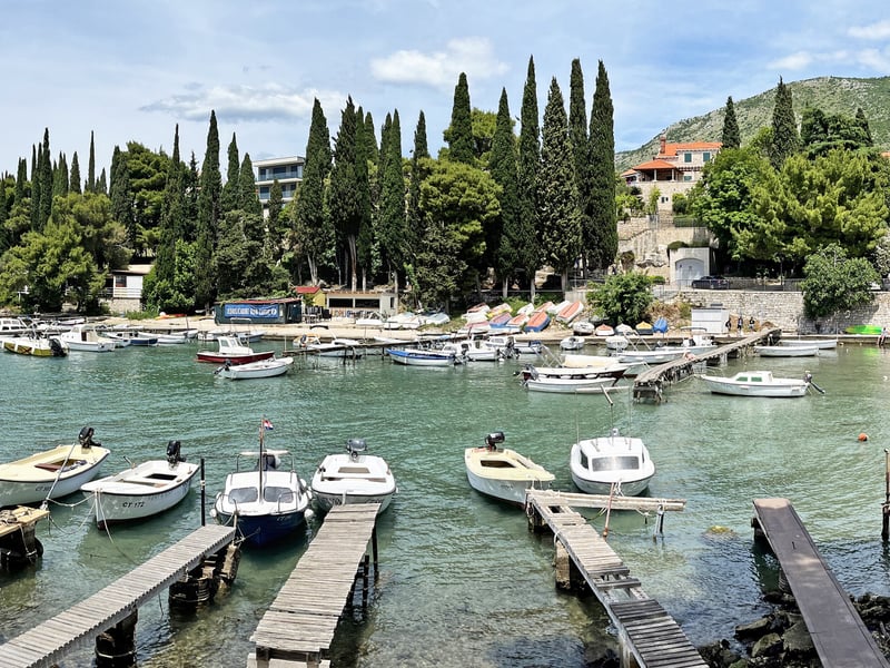 Small boats moored at wooden jetties in Cavtat harbour, with cypress trees and houses behind